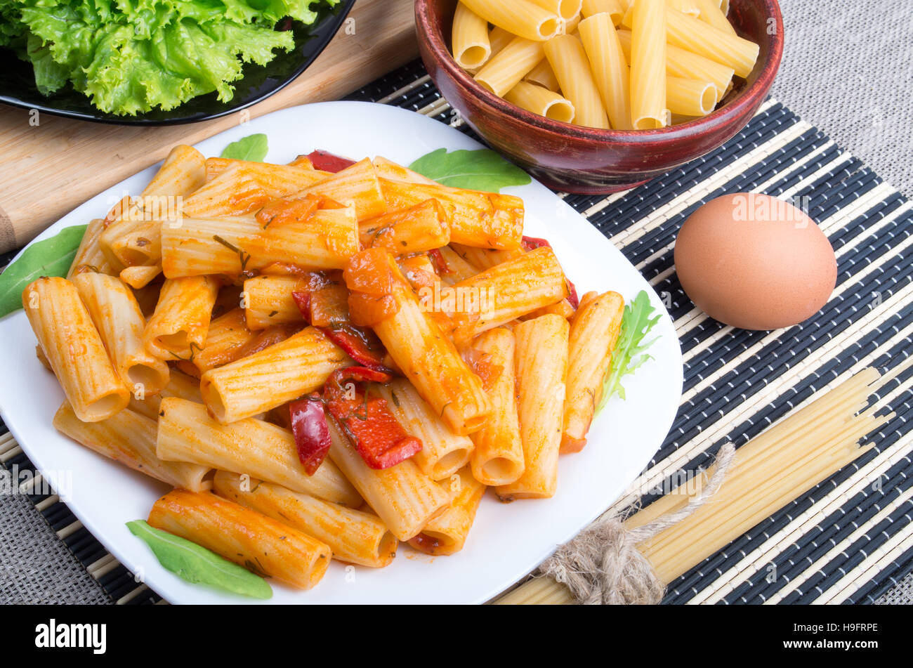Draufsicht auf einen Teller Rigatoni-Nudeln mit Gemüse-Sauce, zusammen mit einem Teller mit Salat und Zutaten zum Kochen Stockfoto