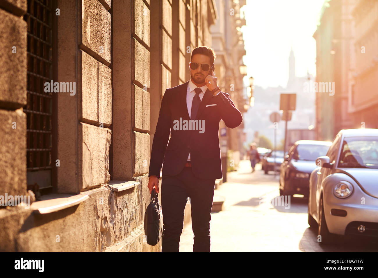 Ein hübscher junger Geschäftsmann auf seinem Handy zu sprechen und zu Fuß auf der Straße in den Sonnenuntergang Stockfoto