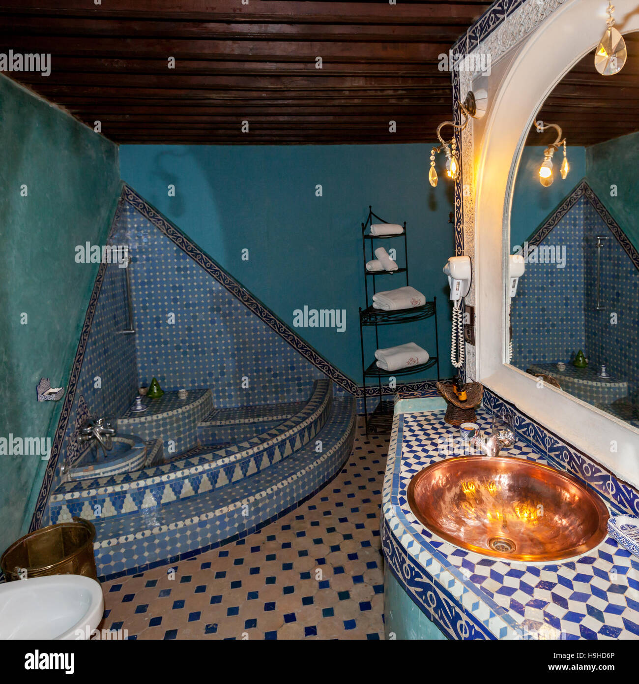 Ein rundes Becken aus poliertem Kupfer bildet das Waschbecken im Badezimmer der Royal Suite des Riad Arabesque in Fès, Marokko, blaue Zellige-Fliesen bedecken die Wände und die erhöhte, traditionell marokkanische Badewanne. Stockfoto