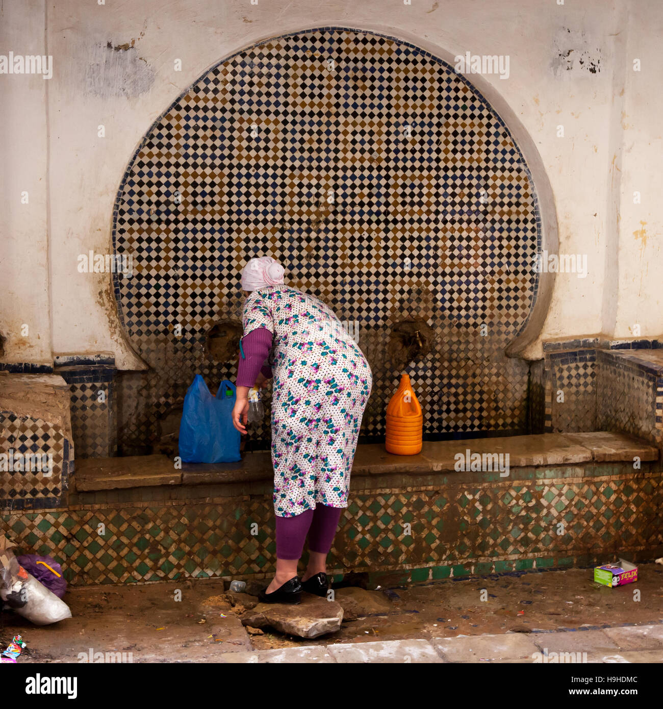 Eine Frau füllt Flaschen und Kanister mit Wasser an einem reich mit Mosaiken verzierten Brunnen in der Medina von Fès, Marokko. Eine blaue Einkaufstasche und ein orangefarbener Kanister stehen am Rand des Beckens bereit. Stockfoto