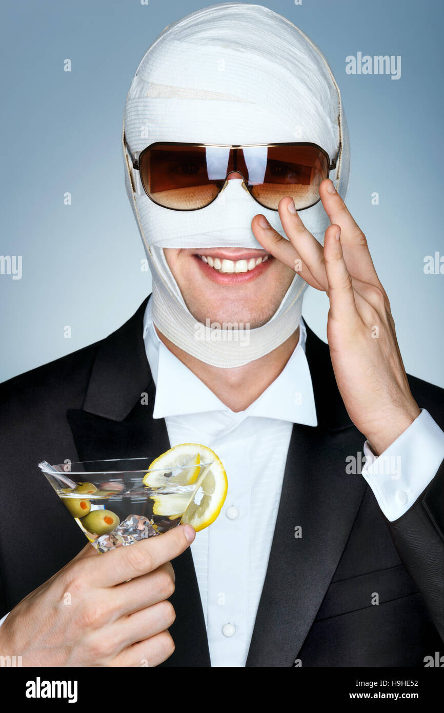 Schönheit-Opfer mit der Binde auf seinem Gesicht nach Facelift. Glamouröse Mann mit Sonnenbrille und mit Glas Martini cocktail. Plastische Chirurgie-Konzept. Stockfoto