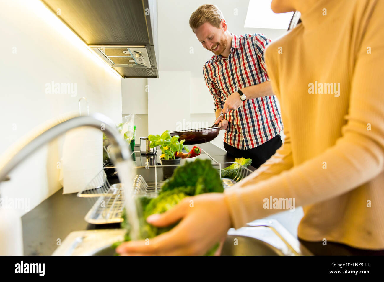 Junge Frau wäscht Grünkohl auf dem Hahn während ein junger Mann in den Hintergrund-Kochen Stockfoto