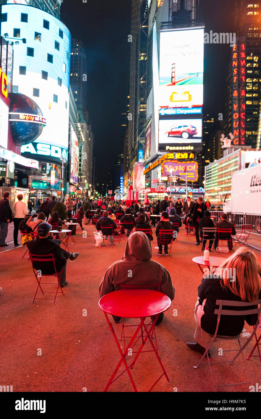 Eine Gruppe von Menschen sitzen auf Stühlen auf dem Gehweg mit dem Rücken zur Kamera beobachten die großen Bildschirme manchmal eckig, New York City, in der Nacht. Stockfoto