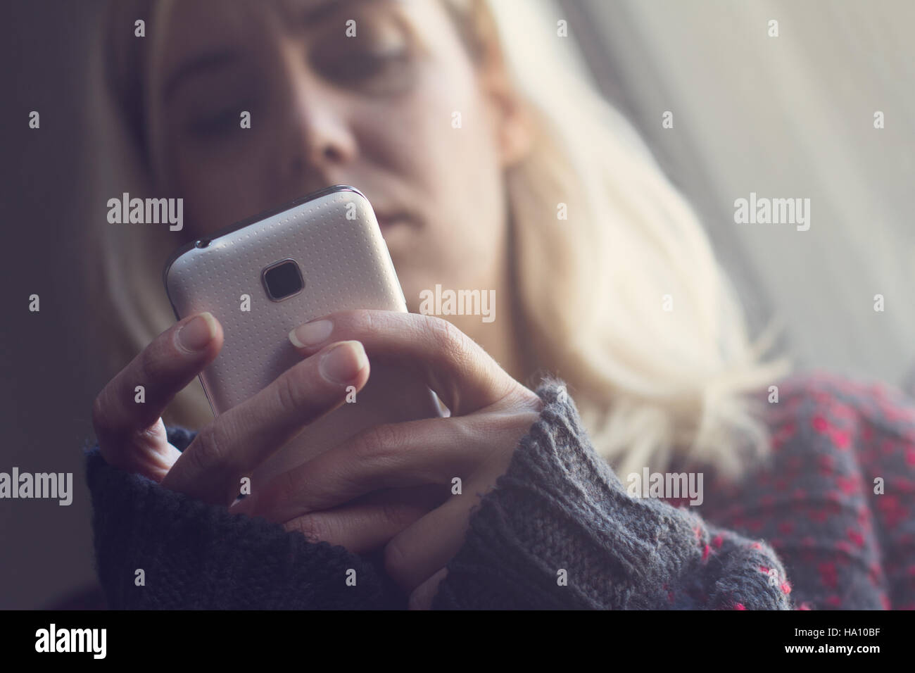 Vorderansicht der blonden Frau mit langen Haaren und dunklen Pullover hält eine weiße Telefon im Morgenlicht Stockfoto