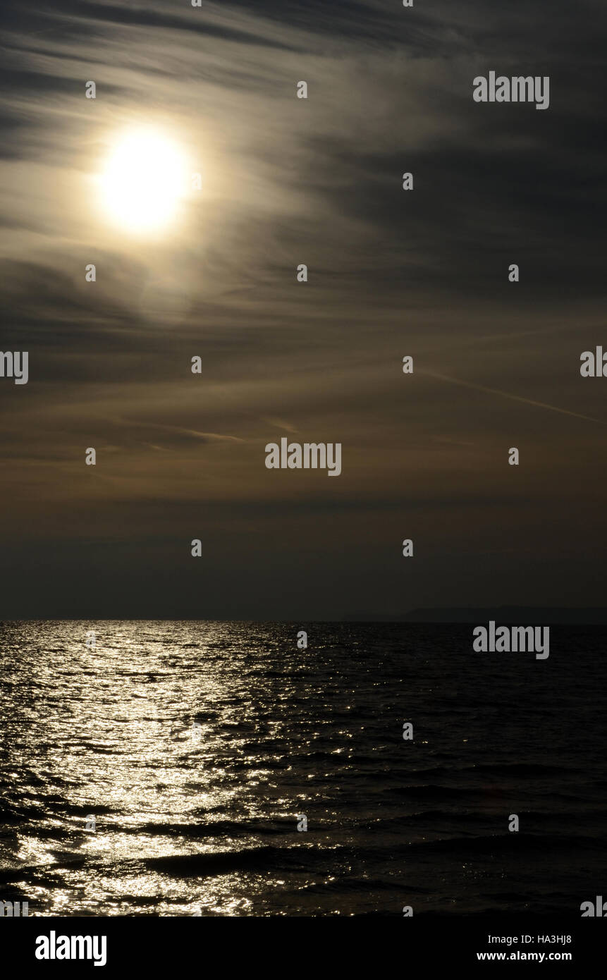 Die Sonne wird gesehen durch einen dünnen Schleier von Wolken, über das Meer, die Szene wird aufgrund der Hintergrundbeleuchtung dunkel. Stockfoto