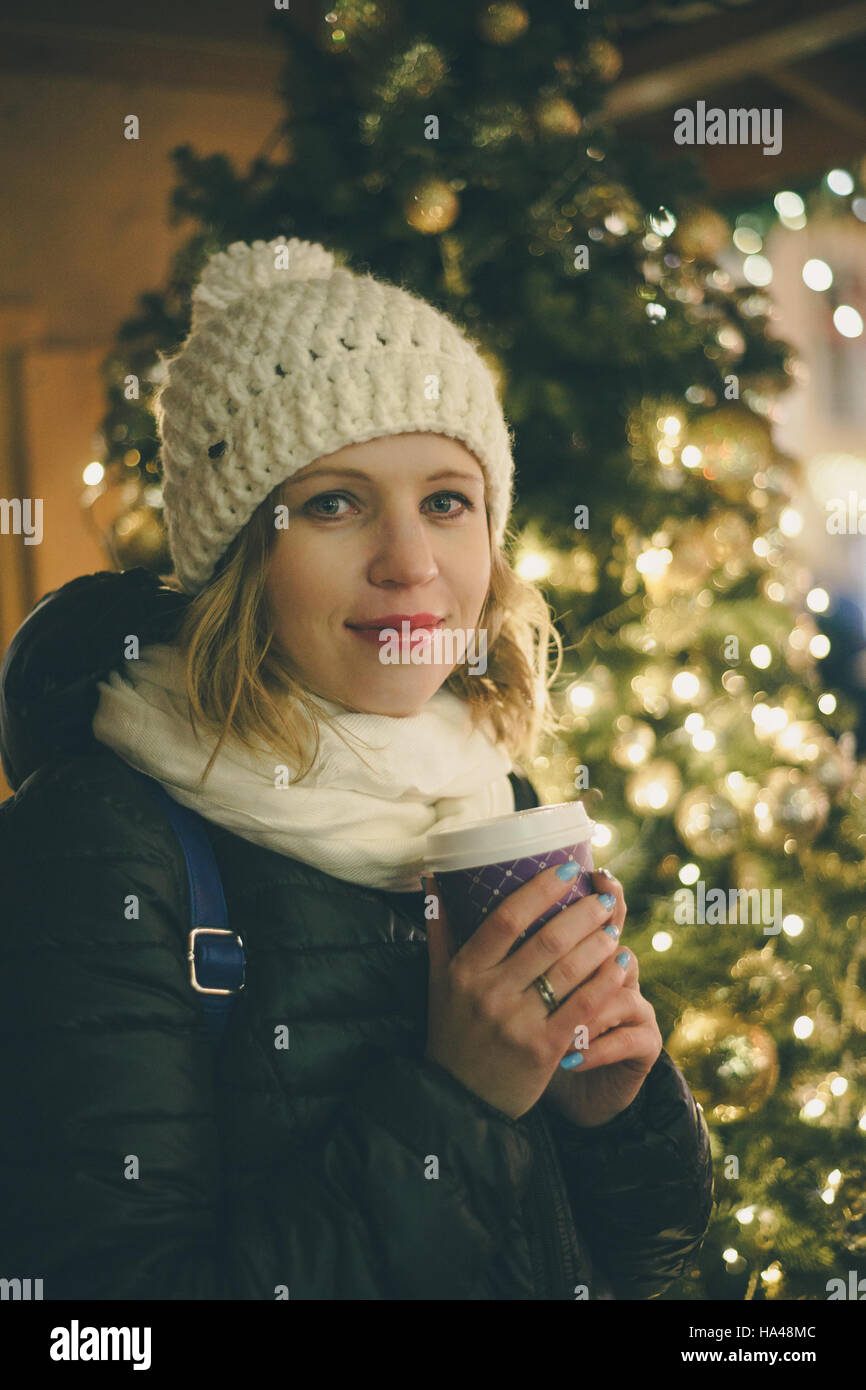 Lächeln Sie, junge Dame mit Tasse Heißgetränk auf Weihnachtsmarkt in der Nähe von Weihnachtsbaum. Winter Urlaub Konzept Stockfoto
