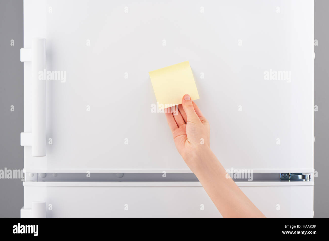 Weibliche Hand mit hellen gelben klebrigen Blankopapier Hinweis auf weißen Kühlschranktür Stockfoto