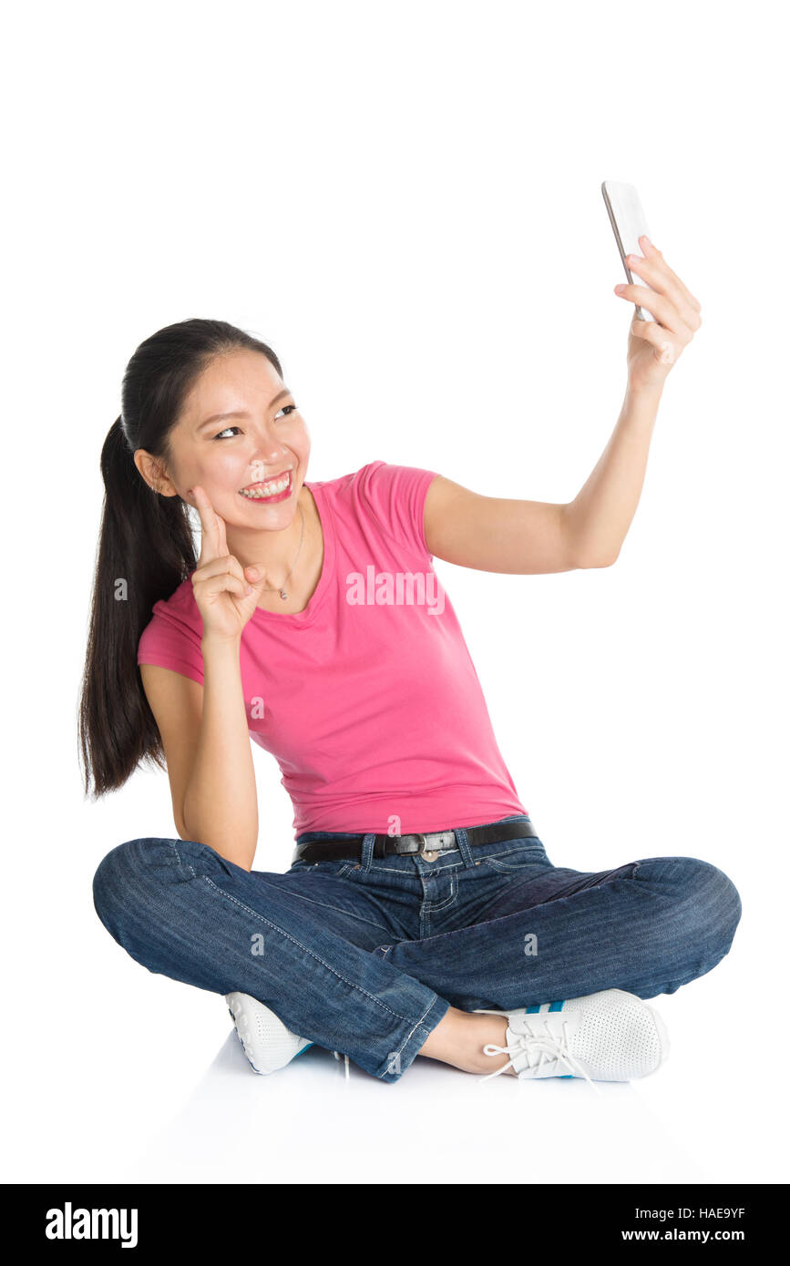 Ganzkörper junge asiatische Mädchen in rosa Hemd nehmen selbst Foto oder Selfie mit Smartphone, sitzen am Boden, voller Länge isoliert auf weißem Hintergrund. Stockfoto