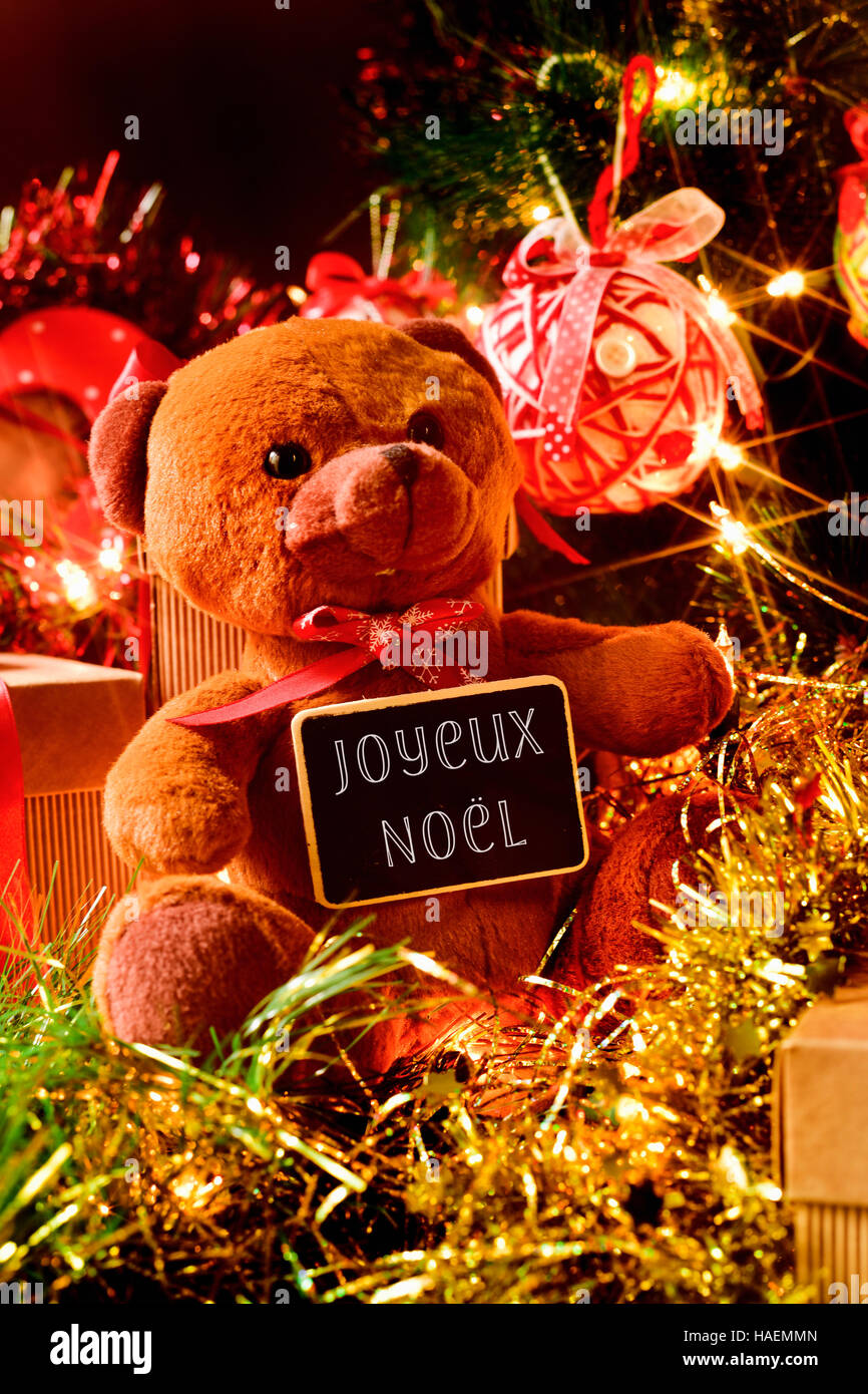 Nahaufnahme eines Teddybären mit einer Tafel mit dem Text Joyeux Noel, Frohe Weihnachten, geschrieben Französisch, und einige Geschenke unter dem Weihnachtsbaum Ornament Stockfoto