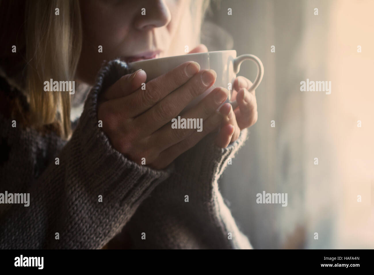 Frau im grauen Strickpullover mit traditionellen Motiven, die eine weiße Tasse heißen Kaffee zu trinken, mit Dampf kommt im Licht morgens Stockfoto