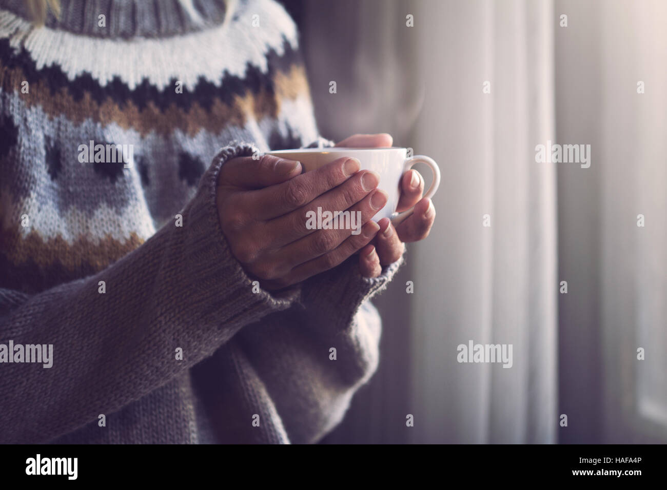 Frau im grauen Strickpullover mit traditionellen Motiven in Händen hält, eine weiße Tasse heißen Kaffee mit Dampf kommt im Licht morgens Stockfoto