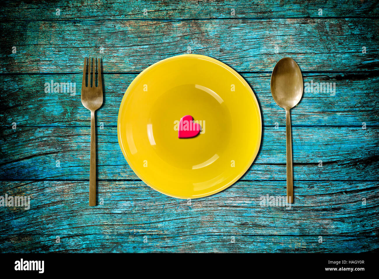rotes Herzform auf gelben leeren Teller mit Gabel und Löffel auf blauen Vintage Holztisch für Liebe-Abendessen-Konzept Stockfoto