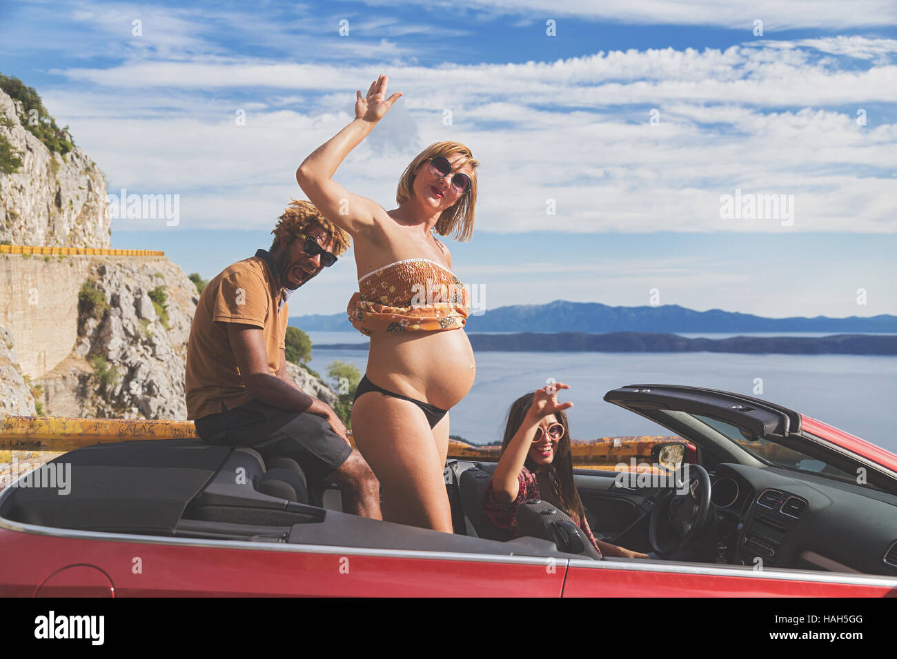 Gruppe der glückliche junge Menschen im roten Cabrio. Schwangere Frau zeigt ihren Bauch. Matte Filter angewendet. Stockfoto
