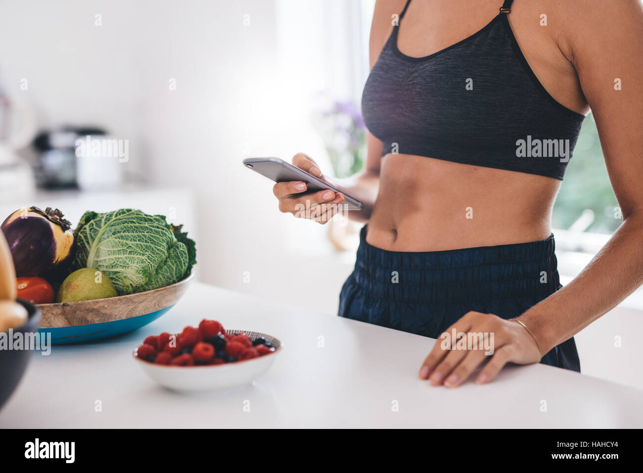 Aufnahme der jungen Frau mit Handy in der Küche mit Obst und Gemüse auf Küchentisch beschnitten. Stockfoto