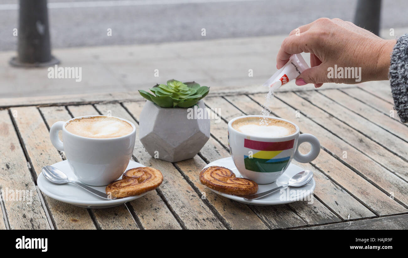 Zwei Tassen Kaffee auf einem Holztisch im Freien mit dänischen Keksen und Löffel Barrista. Frau Gießen Zucker in Stockfoto