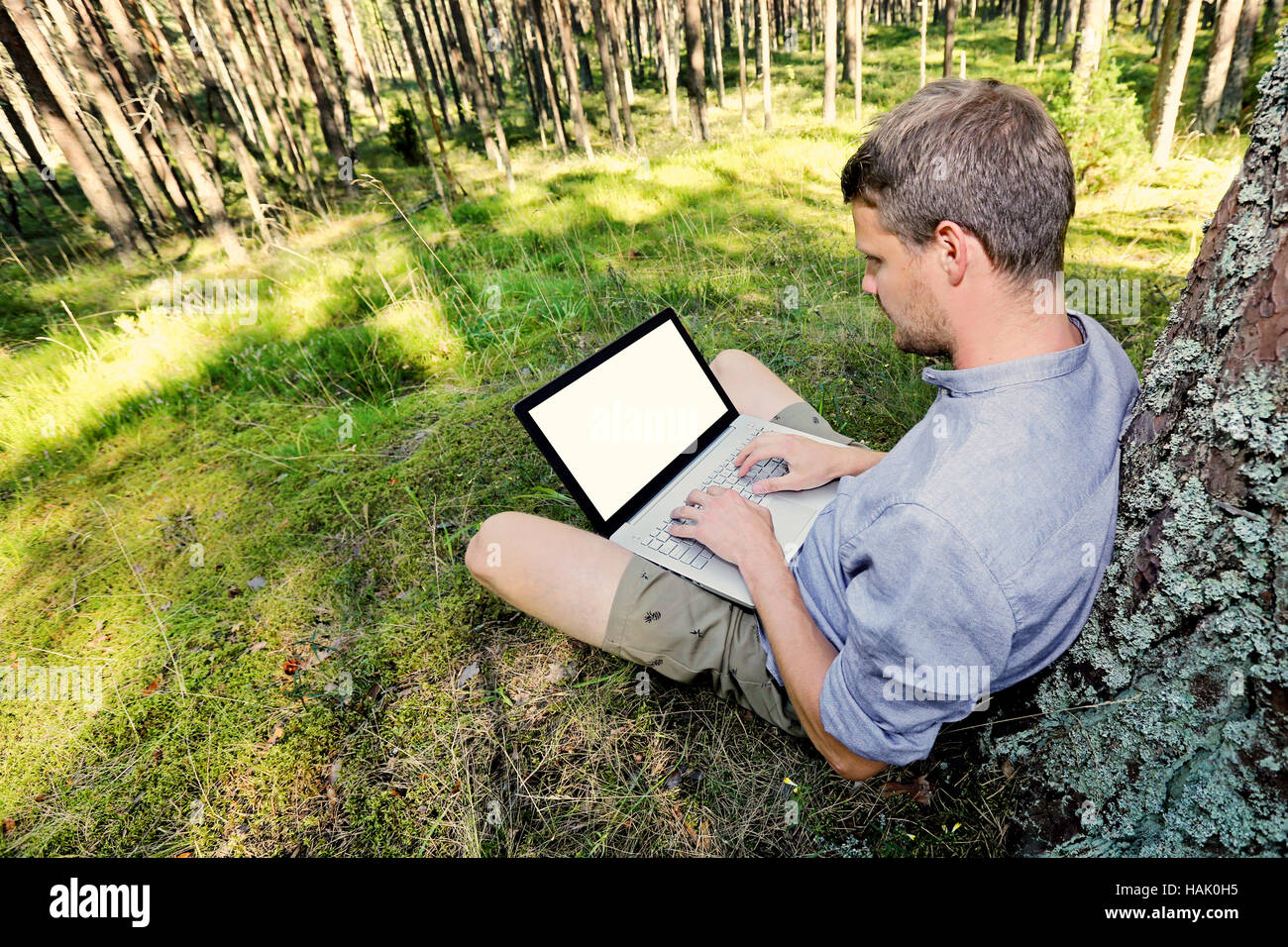Mann sitzt mit seinem Laptop arbeitet gegen einen Baum im Wald, Stockfoto