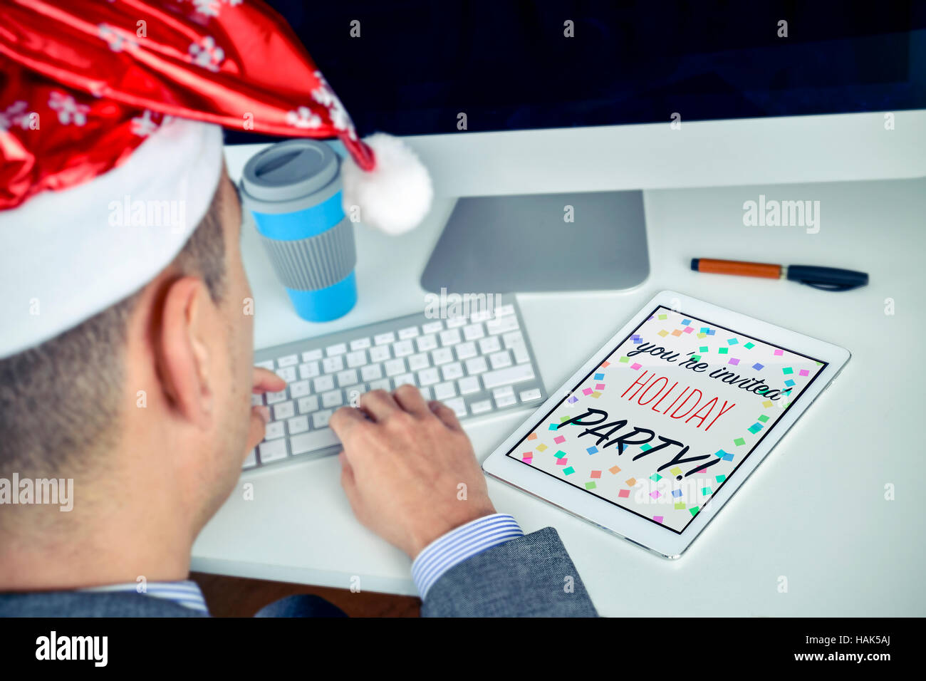 kaukasischen Jüngling mit einem Santa Hut sitzt an seinem Schreibtisch auf seinem Tablet, wo Sie die Text-Weihnachts-Party lesen können, Sie sind herzlich eingeladen Stockfoto