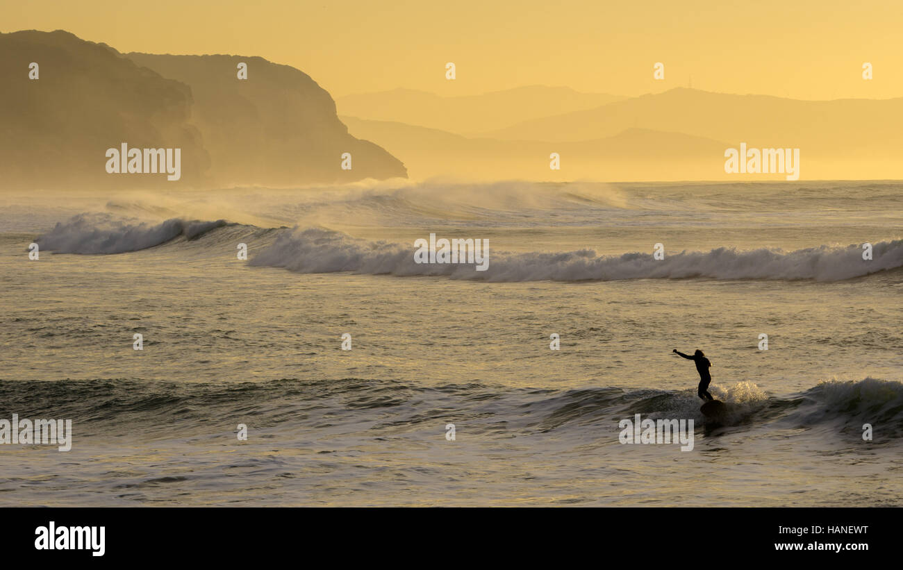 Ein Mann mit seinem Longboard Surfen die Wellen in der Caños de Meca bei Sonnenaufgang, im Hintergrund Actitilados aus Sandstein Stockfoto