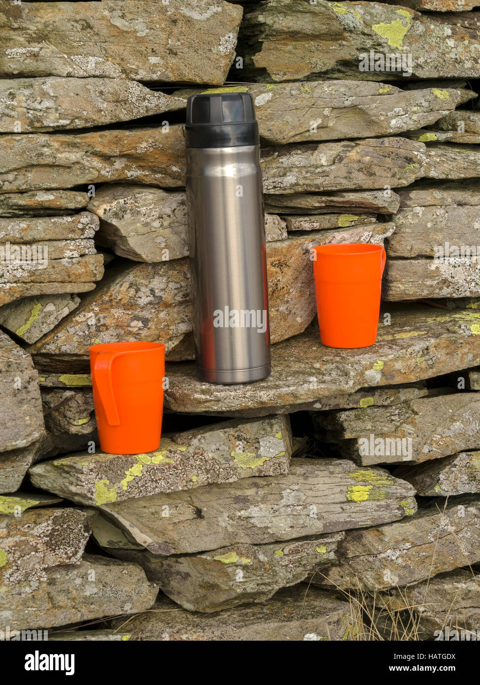 Thermoskolben aus Edelstahl und zwei orangefarbene Kunststoffbecher an trockenen Steinwänden. Stockfoto