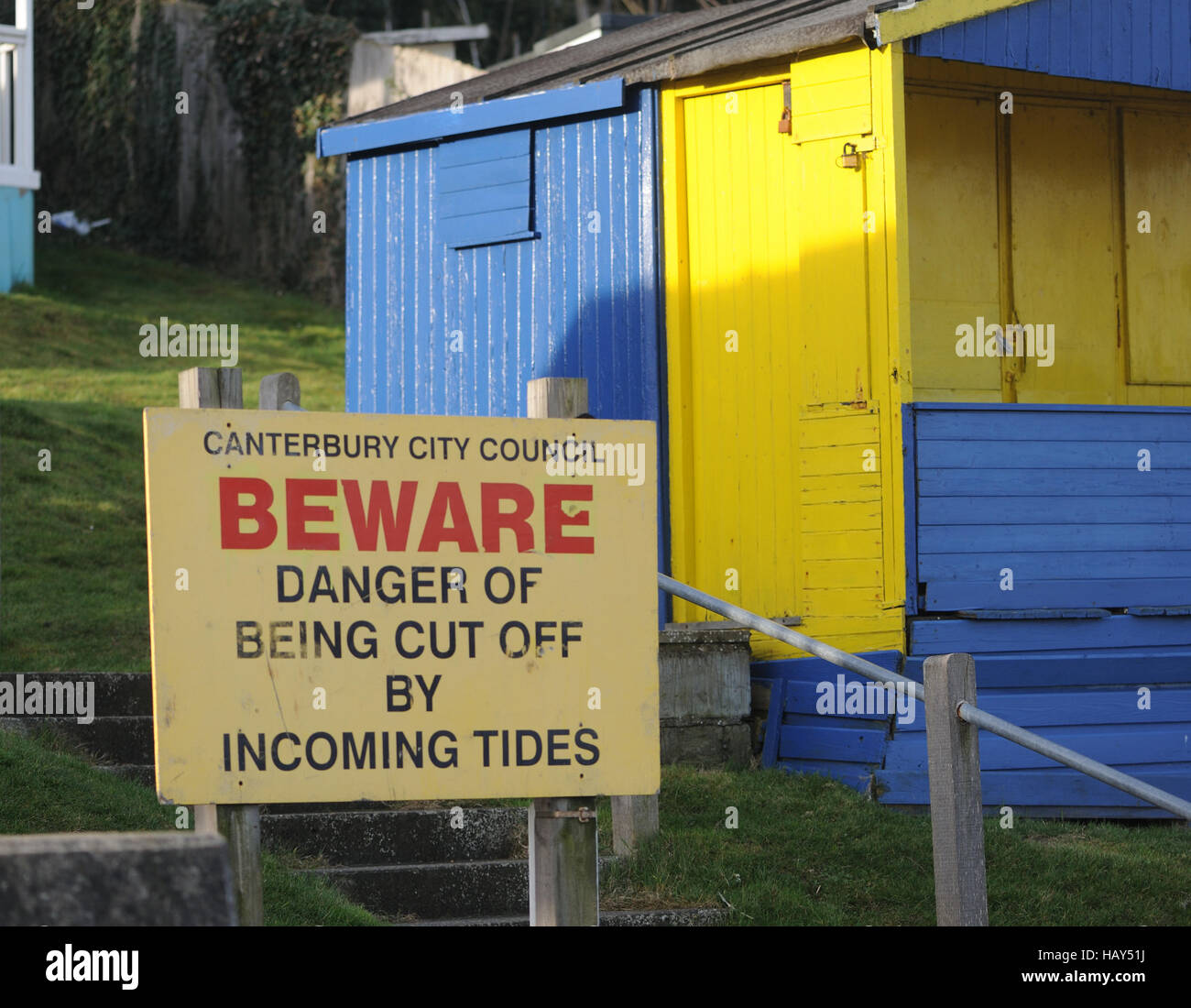 Eine Schild Warnung über sein Abschneiden von ankommenden Gezeiten vor eine blaue und gelbe Strandhütte am Strand von Whitstable Stockfoto