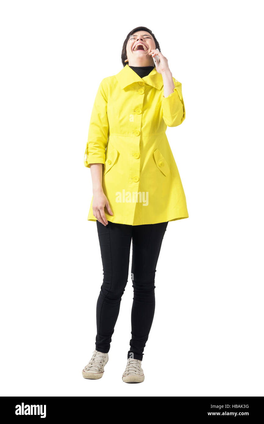 Glückliche junge kurzhaarige Frau im gelben Mantel laut lachend auf Ihrem Handy. Ganzkörper-Länge-Porträt isoliert auf weiss Stockfoto