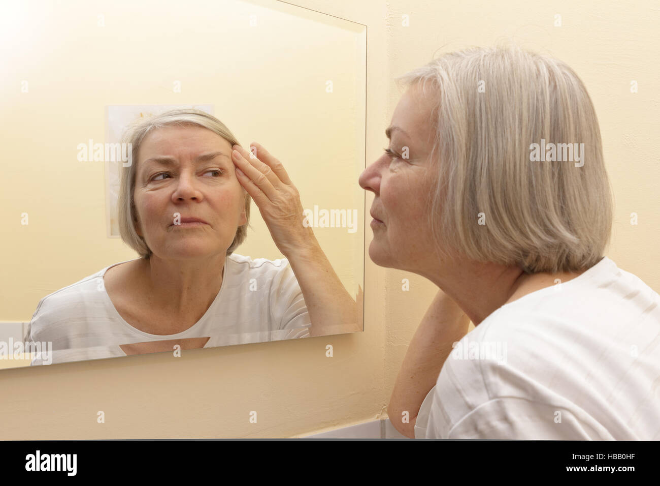 Ältere Frau mit dem grauen Haar ihre Gesichtshaut Prüfung im Spiegel ihrer gelben Badezimmer, altern Konzept Stockfoto