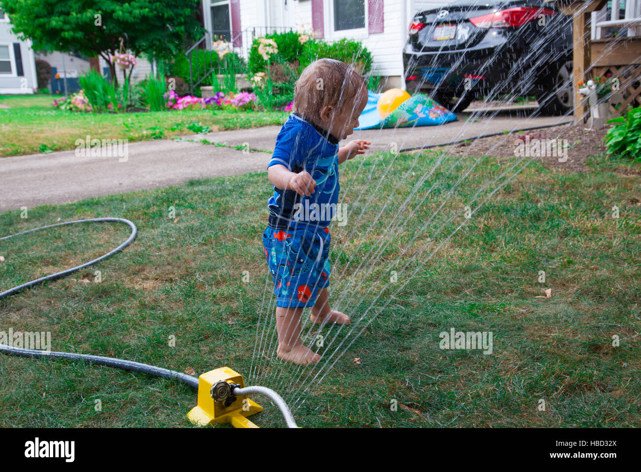 Kaukasische Baby Junge spielt mit einem Wasser-Sprenger in seinem vorderen Rasen im Sommer Stockfoto