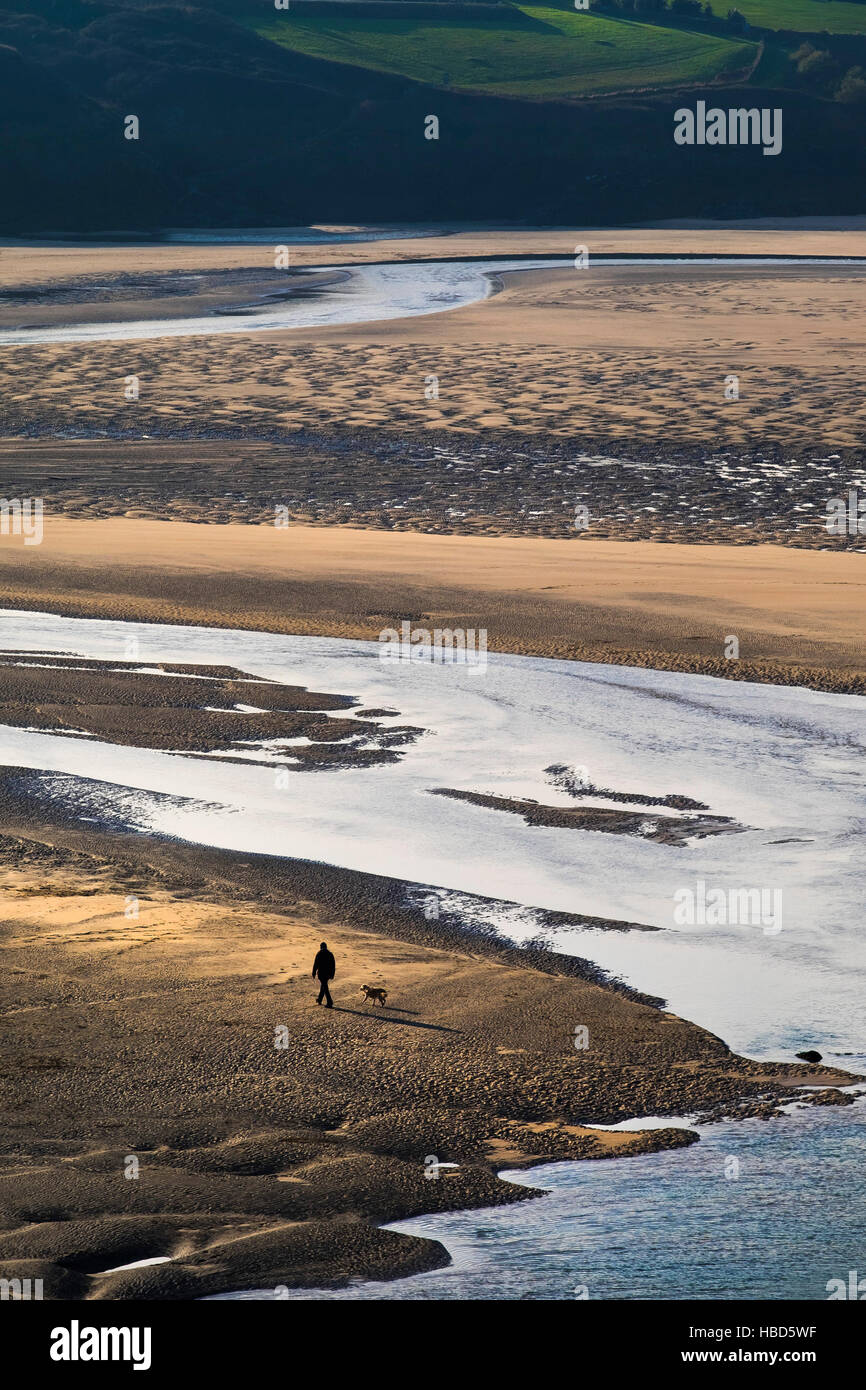 Eine Dogwalker und seinem Hund gesehen in der Silhouette und aus der Ferne auf dem preisgekrönten Crantock Beach in Newquay, Cornwall. Stockfoto