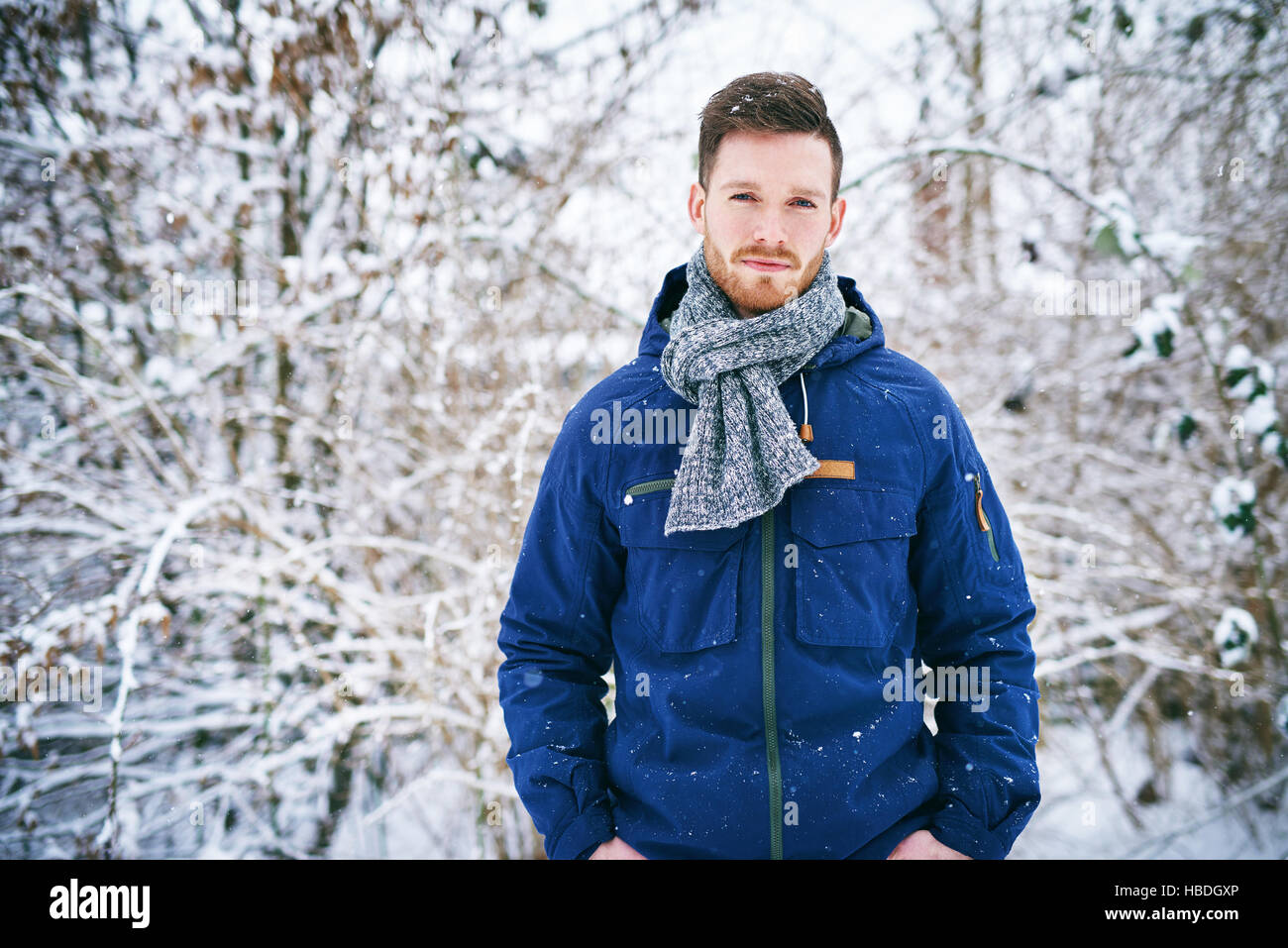 Junge bärtige Mann mit warmen Mantel mit Händen in den Taschen, Blick in die Kamera gegen schneebedeckten Baum. Stockfoto