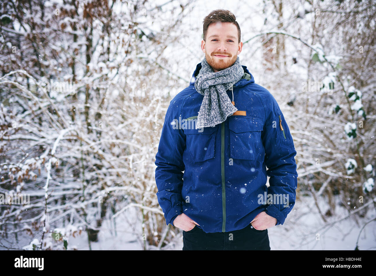 Jung, lächelnd bärtiger Mann in warmen Mantel Blick in die Kamera auf unscharfen Hintergrund der verschneiten Wald. Stockfoto