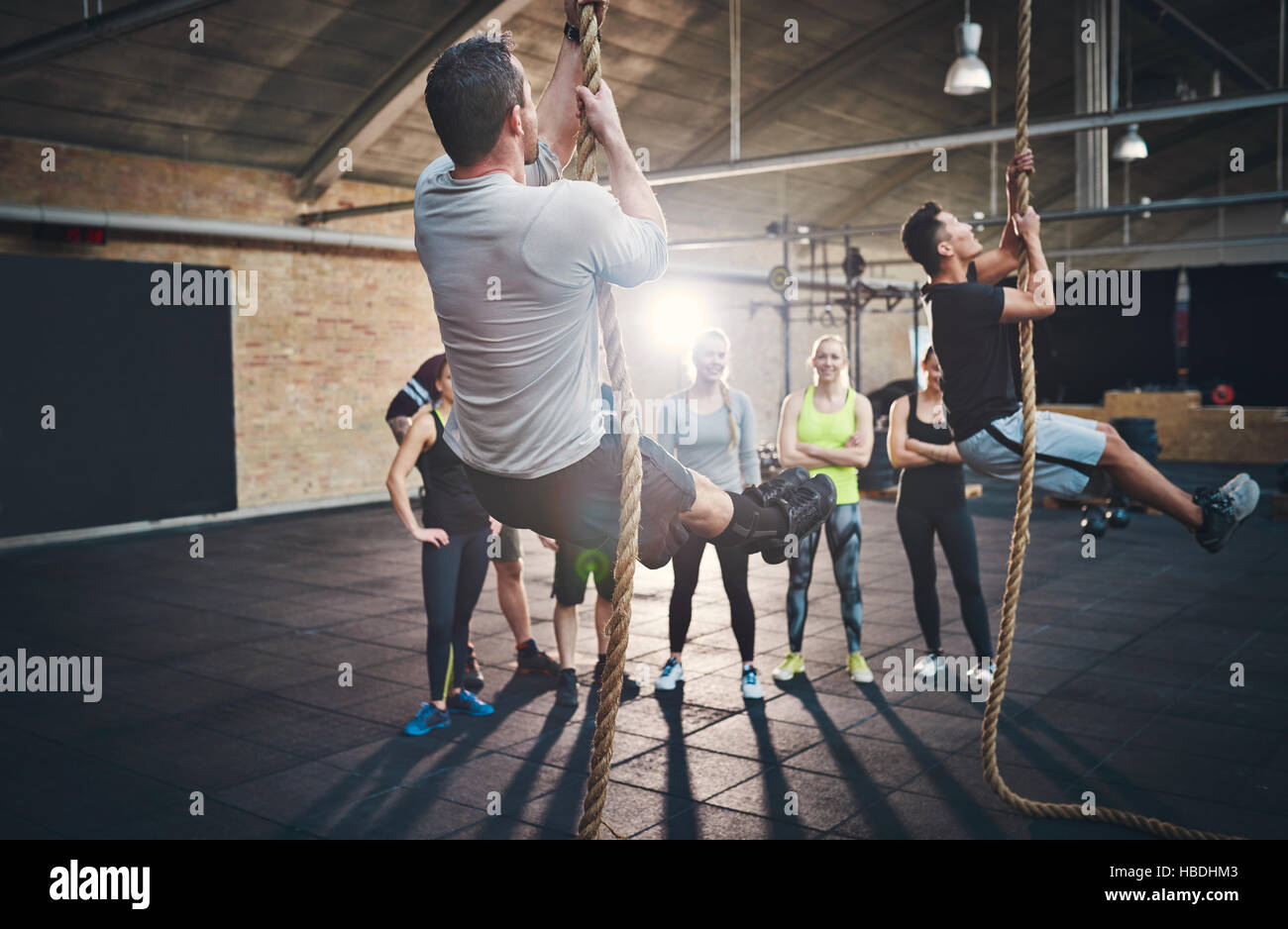 Zwei Männer in kurzen Hosen Dicke Klettertau während Fitness-Übungen in Cross-Fit Training mit anderen Erwachsenen beobachtete sie von hinten Stockfoto