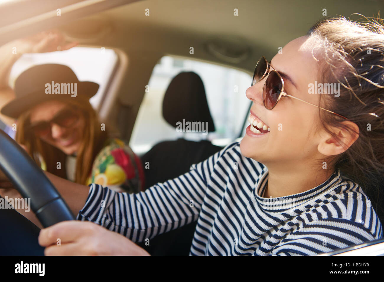 Attraktive moderne, junge Frau in Sonnenbrille lachte, als sie ein Auto in der Stadt Spaß mit ihren weiblichen Passagier Laufwerke, bis Ansicht schließen durch das offene Stockfoto