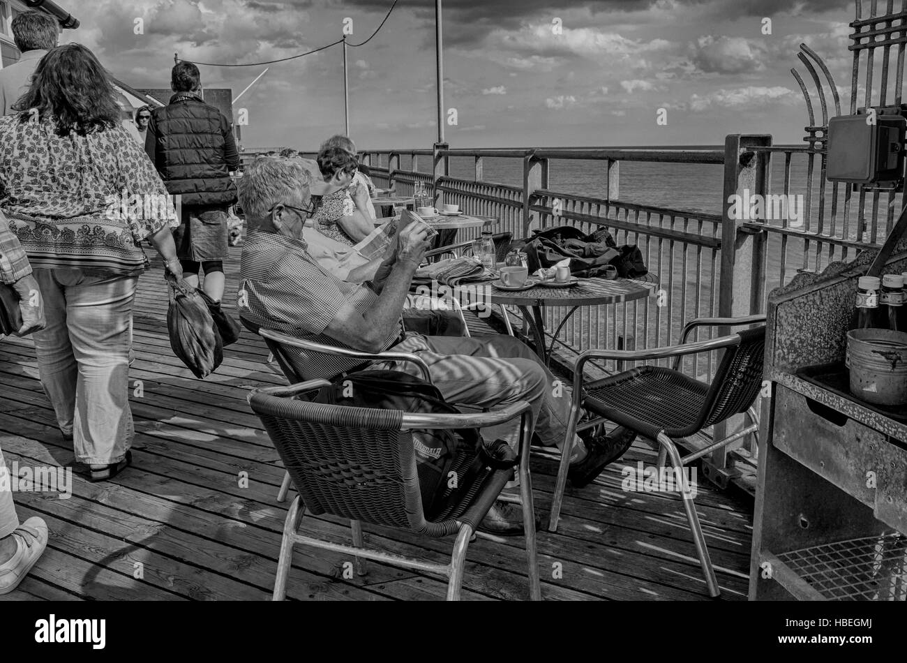 Southwold, England. Ältere Menschen genießen das sonnige Wetter entlang Southwold pier Tee trinken und Zeitung lesen. Die jüngeren Besucher sind zu Fuß durch. Foto umgewandelt, HDR, High Dynamic Range, für ein dramatischer kontrastreiches Bild. Stockfoto