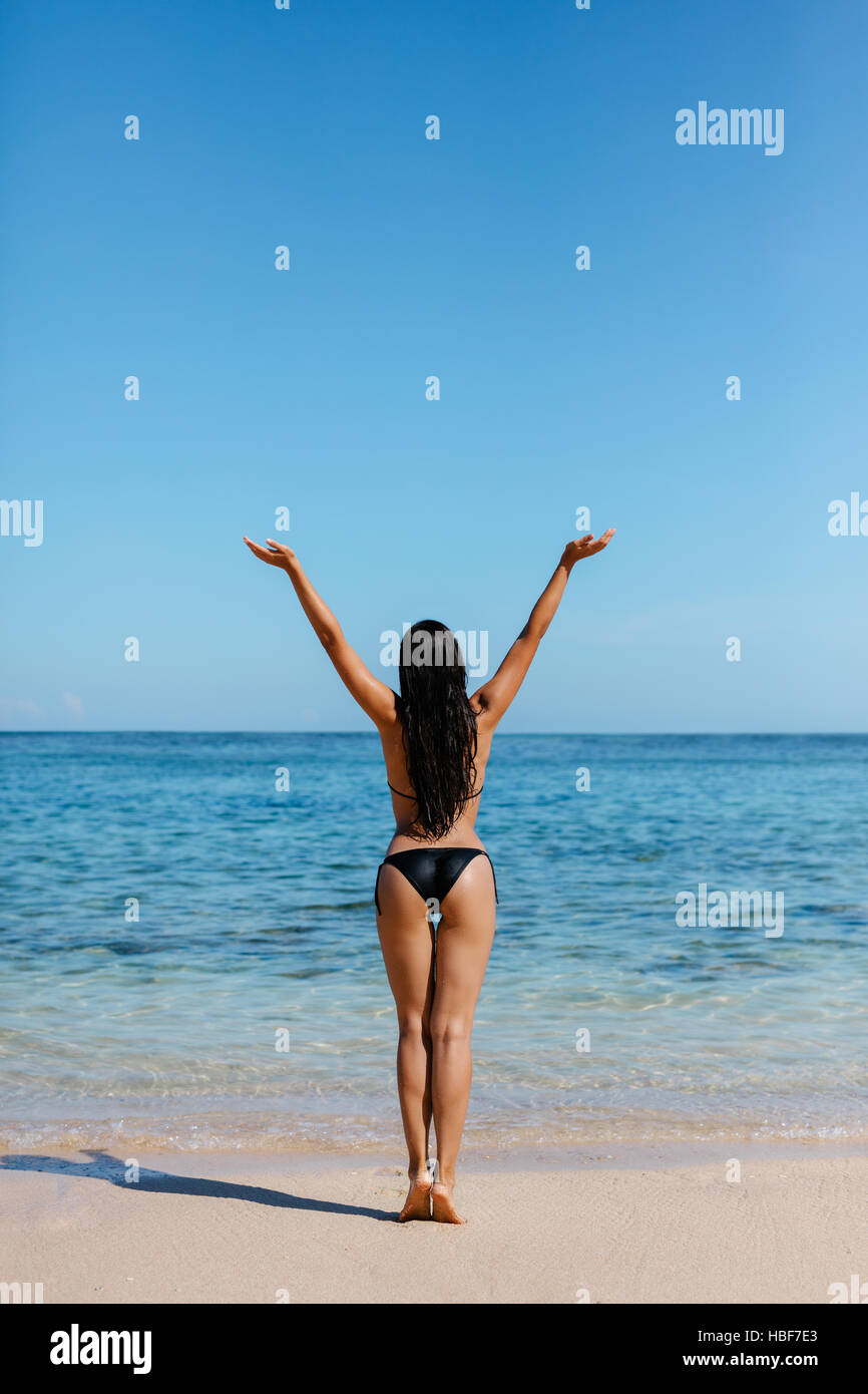 Rückansicht des jungen Frau mit Bikini am Strand. Junge Frau im Badeanzug stehen an der Küste mit ihren Händen in Richtung Himmel erhoben. Stockfoto