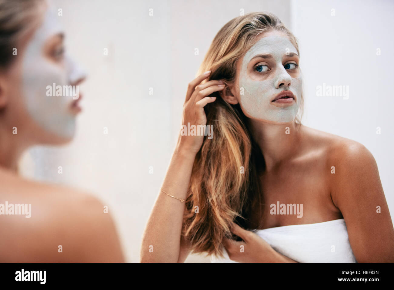 Weibchen mit kosmetische Gesichtsmaske im Badezimmer. Frau in Blick in den Spiegel mit Maske auf ihr Gesicht Handtuch gewickelt. Stockfoto