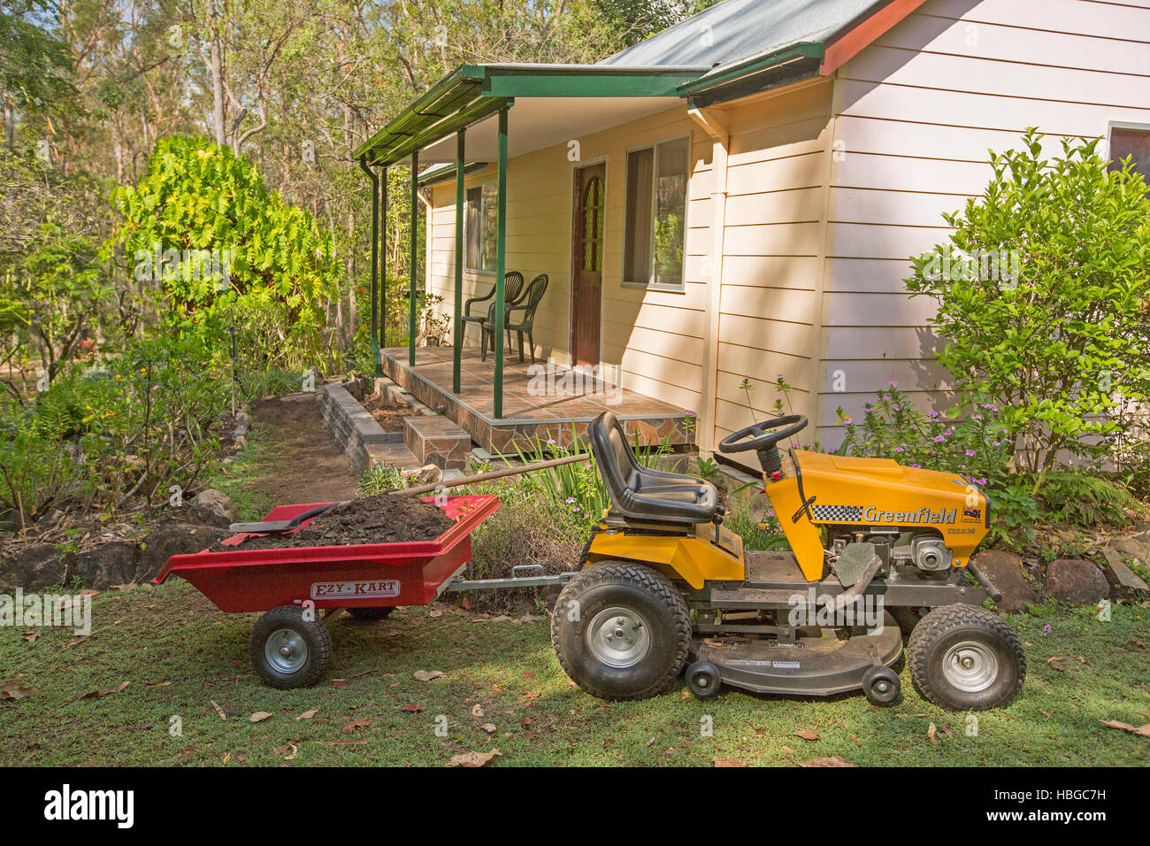 Gelbe Reiten-auf Rasenmäher ziehen kleine rote Anhängers voll des Bodens auf Rasen neben Haus mit Garten von Sträuchern und Bäumen Stockfoto