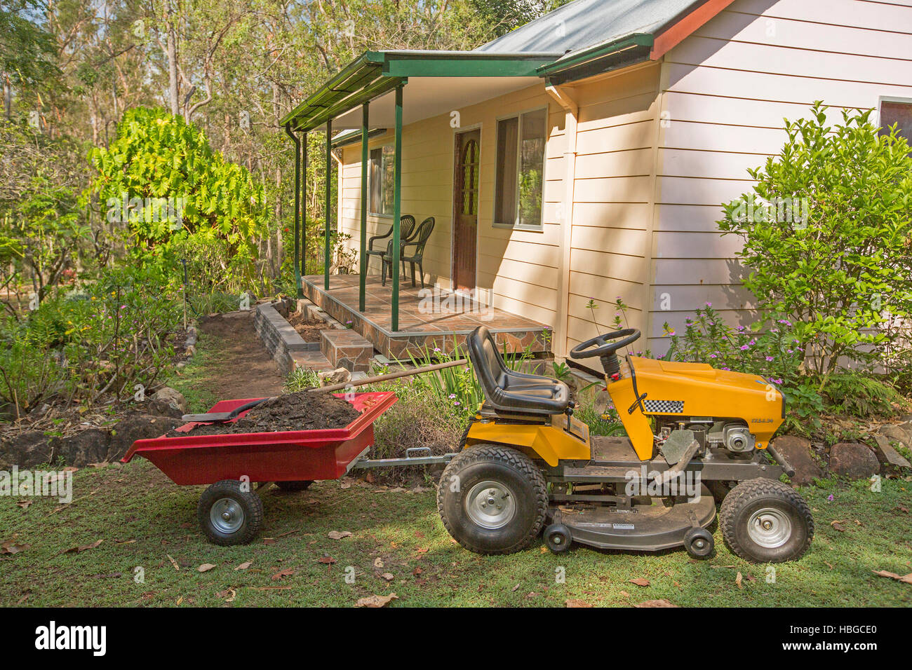 Gelbe Reiten-auf Rasenmäher ziehen kleine rote Anhängers voll des Bodens auf Rasen neben Haus mit Garten von Sträuchern und Bäumen Stockfoto