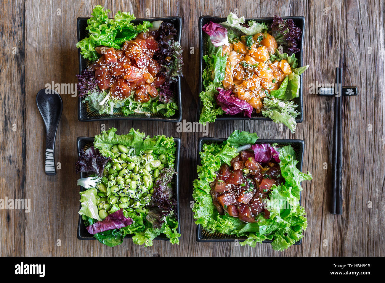 Draufsicht auf 4 Poke-Schüsseln mit Essstäbchen und einem Löffel auf einer Holzfläche. 3 Fischoptionen und eine vegetarische Option. Stockfoto