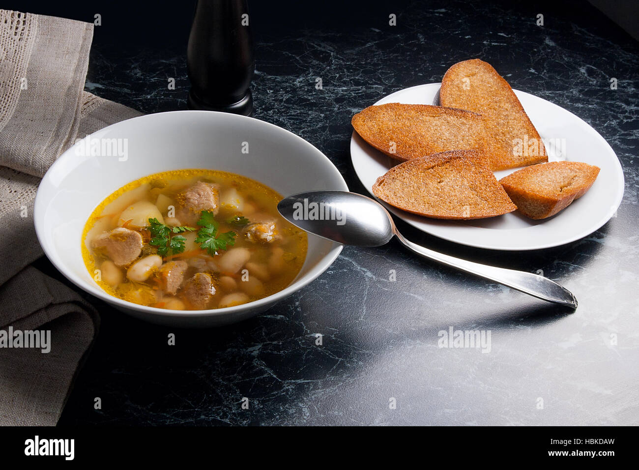 Bohnensuppe in weiße Schüssel mit Metalllöffel auf einem schwarzen Stein Hintergrund. Einige toast auf weißen Teller und schwarze Mühle für Pfeffer auf braunem Tuch. Stockfoto