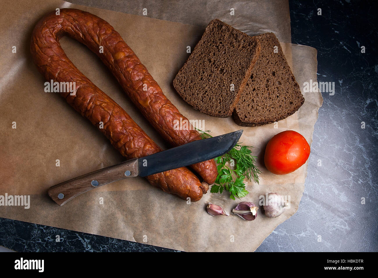 Nahaufnahme Blick geräucherte Wurst mit Petersilie und schwarze Roggenbrot, mehrere Tomaten und Knoblauch auf braunem Packpapier. Vintage Messer auf Wurst. Stockfoto