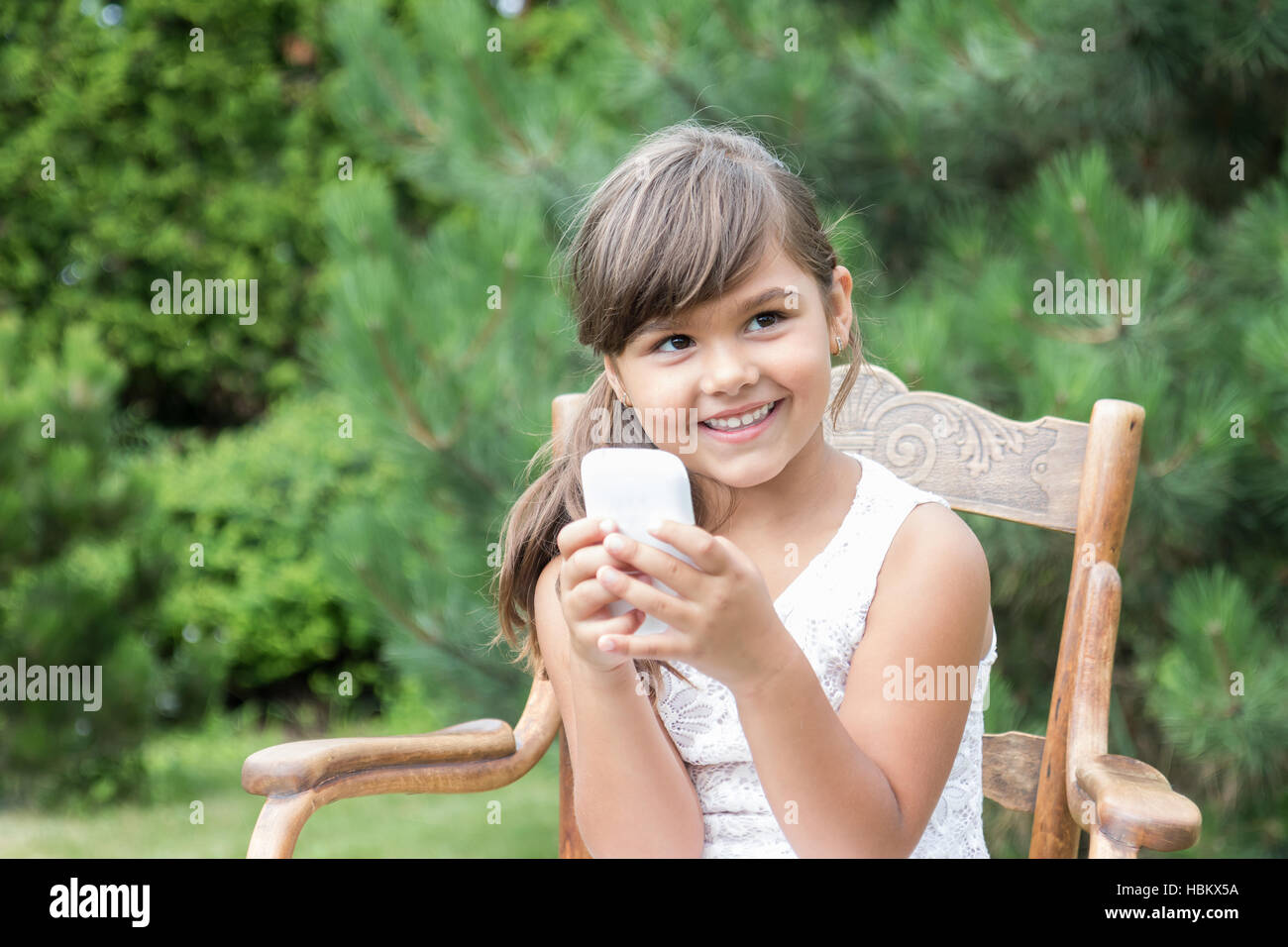 Lächelndes langhaarige Brünette Mädchen sitzt auf und alten Holzstuhl im Freien. Kleines Mädchen hält eine Smartphone in ihre Hände. Stockfoto