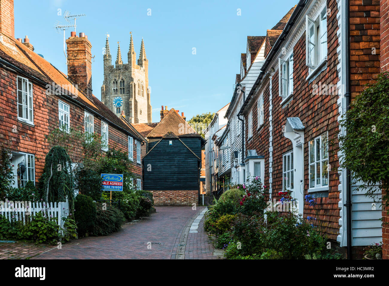 England, Tenterden. Die ix Feld Path' eine schmale gepflasterte Straße, Gehäuse und kleinen Vorgärten auf beiden Seiten. Kirchturm im Hintergrund. Malerisch. Stockfoto