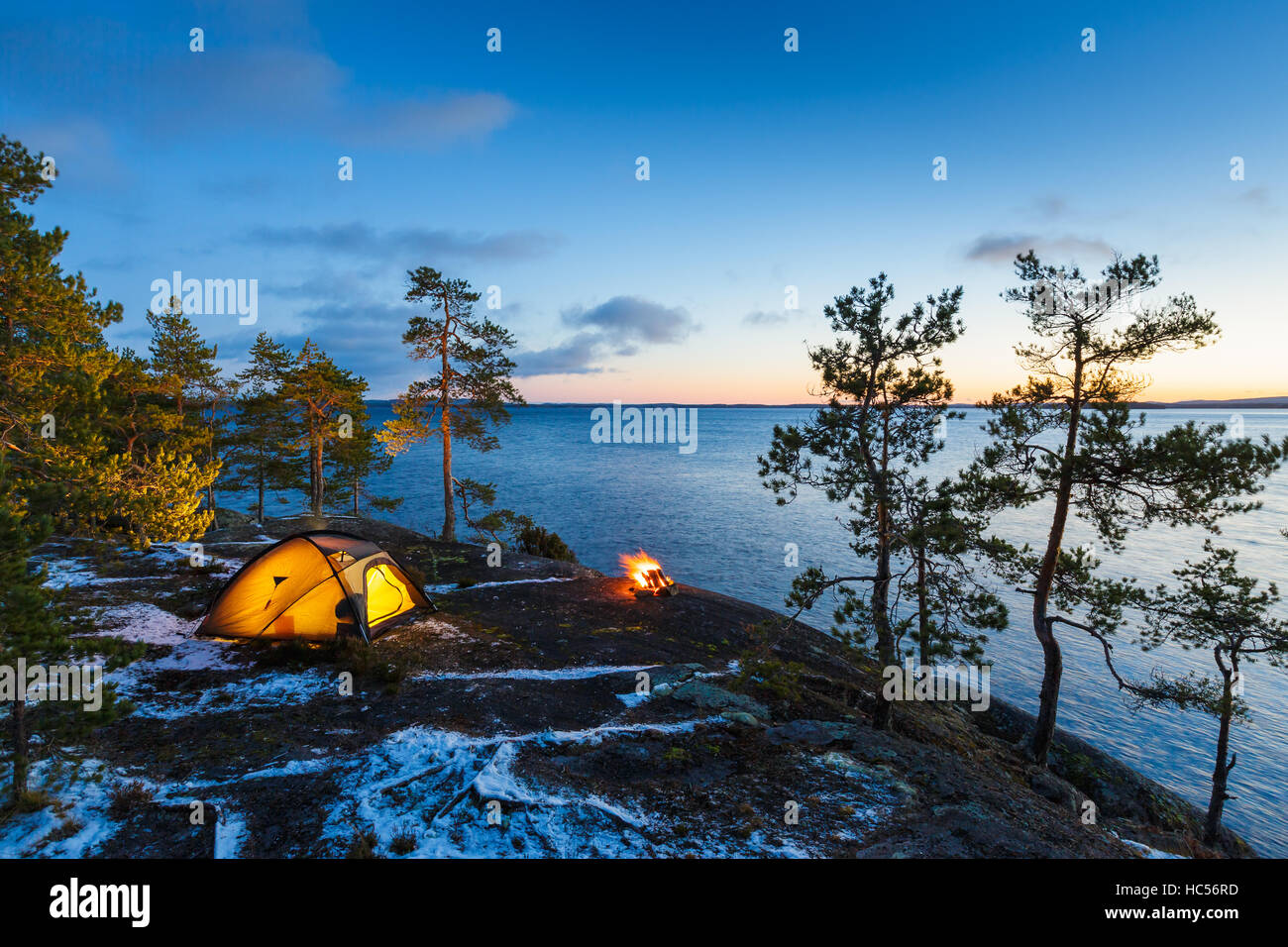 Lagerfeuer und Zelt in der Wildnis am See bei Sonnenuntergang, Schnee auf dem Boden Stockfoto