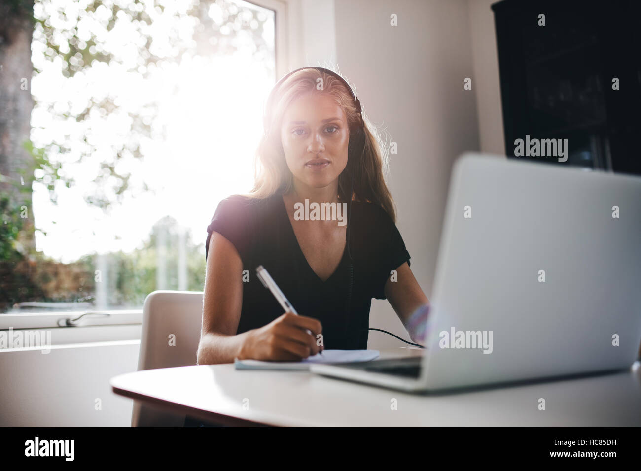 Portrait von junge Frau sitzt in Küche mit Kopfhörern Anmerkung zu schreiben. Frau mit Laptop zu Hause zu studieren. Stockfoto
