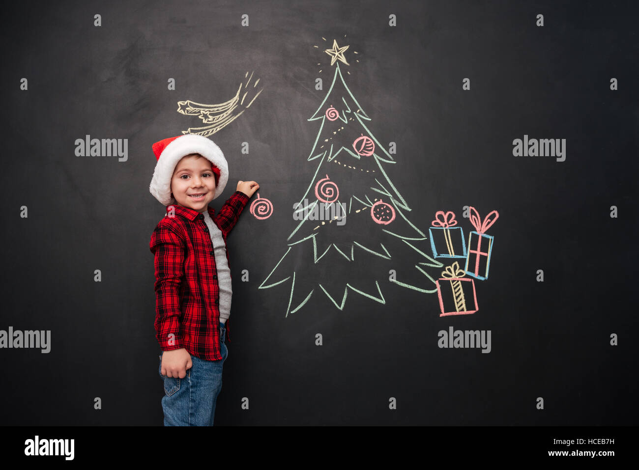 Foto von kleinen niedlichen Kind tragen Hut ziert einen Weihnachtsbaum auf Tafel zeichnen. Blick in die Kamera. Stockfoto