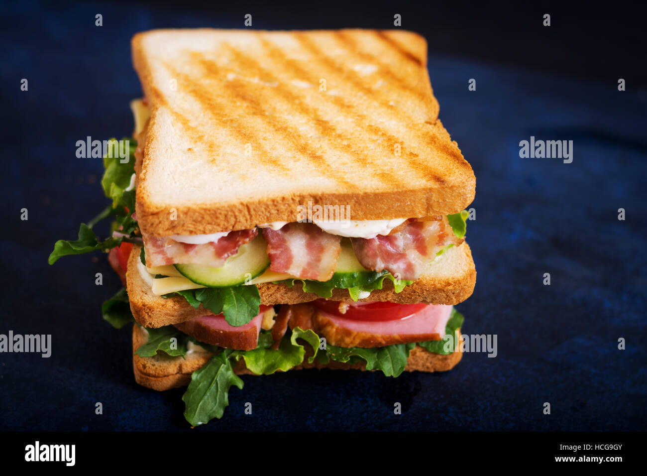 Große Club-Sandwich mit Schinken, Speck, Tomaten, Gurken, Käse, Eiern und Kräutern auf dunklem Hintergrund Stockfoto