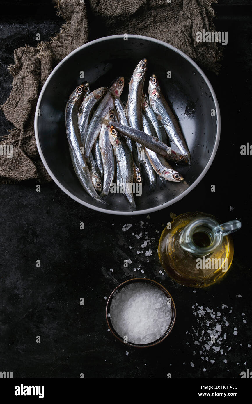 Viele Fische roh frische Sardellen in schwarzen Keramikschale mit Meersalz und Flasche Olivenöl Marinade über schwarzen Metall Hintergrund. Ansicht von oben. Meer Stockfoto
