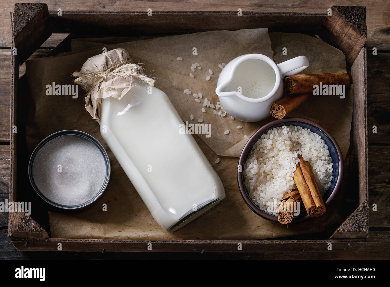 Zutaten für die Herstellung von Milchreis. Schüssel mit ungekochtem Reis, Zucker, Zimtstangen, Flasche Milch und Creme in Holzkiste über alte hölzerne Kanne Stockfoto