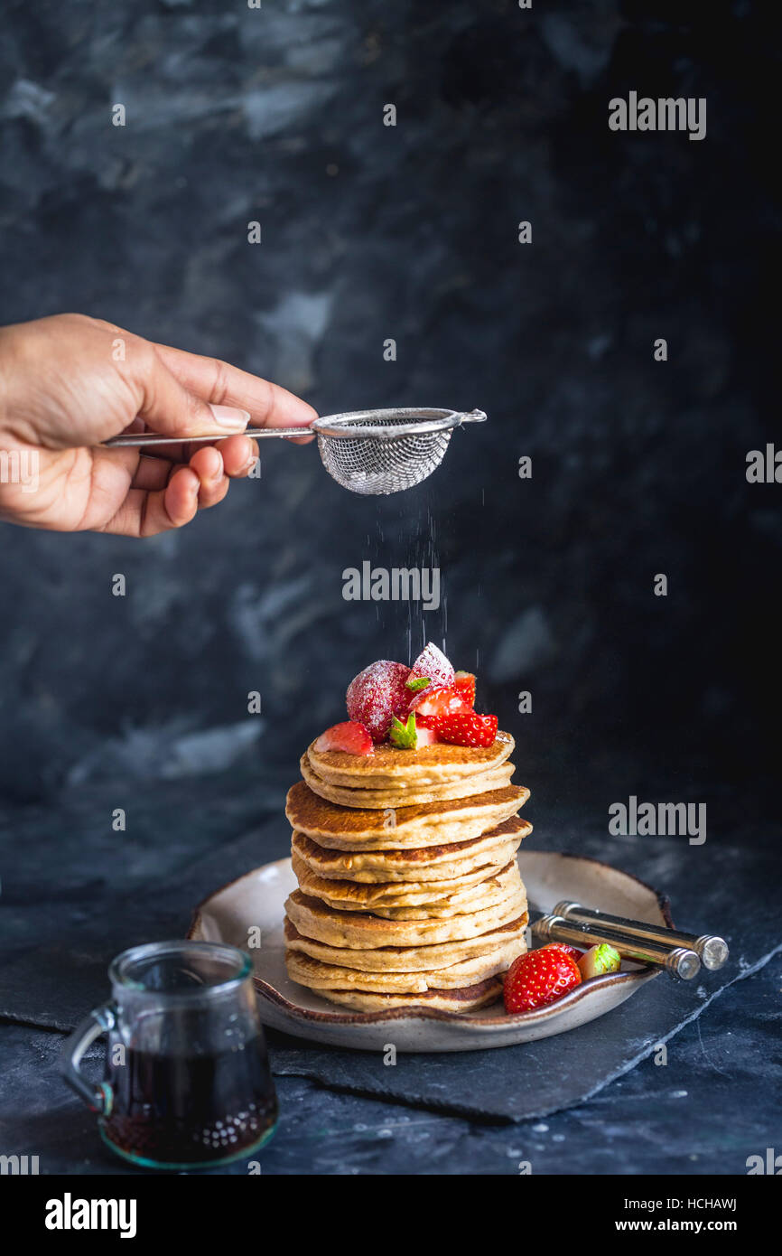 Abstauben Icing Sugar auf Stapel von Gluten freie Hafer Mehl Pfannkuchen serviert mit gehackten Erdbeeren und Ahornsirup Stockfoto