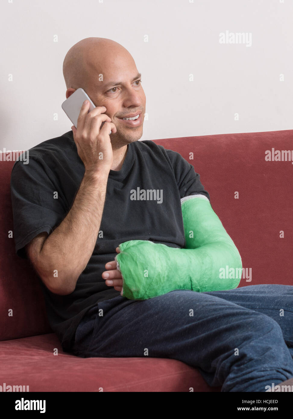 Junger Mann mit einem Arm und Ellenbogen in einem blauen Pflaster / Fiberglas gegossen zu Hause glücklich im Gespräch auf seinem Handy nach den Arm gebrochen hatte Stockfoto